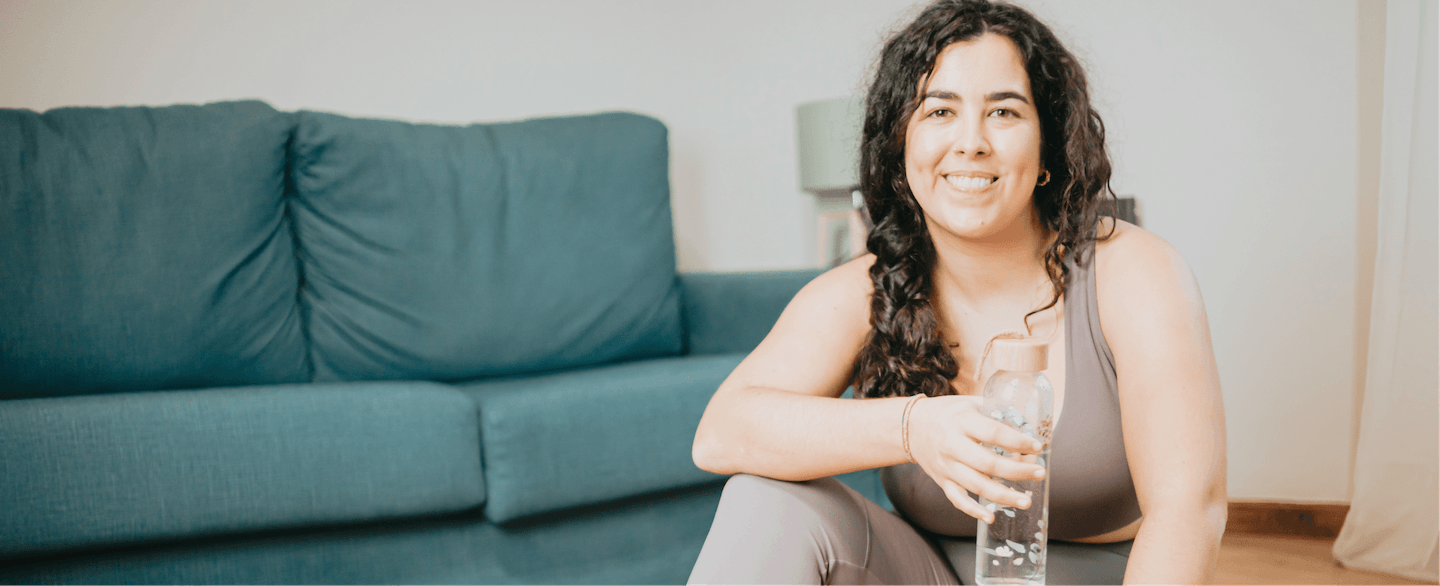 healthy woman in her 30s smiling, sitting on floor, wearing workout clothes and holding water bottle
